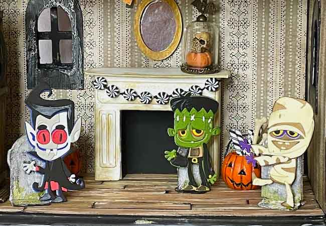 3 paper figures dressing Halloween costumes in a Halloween dollhouse decorated for a party