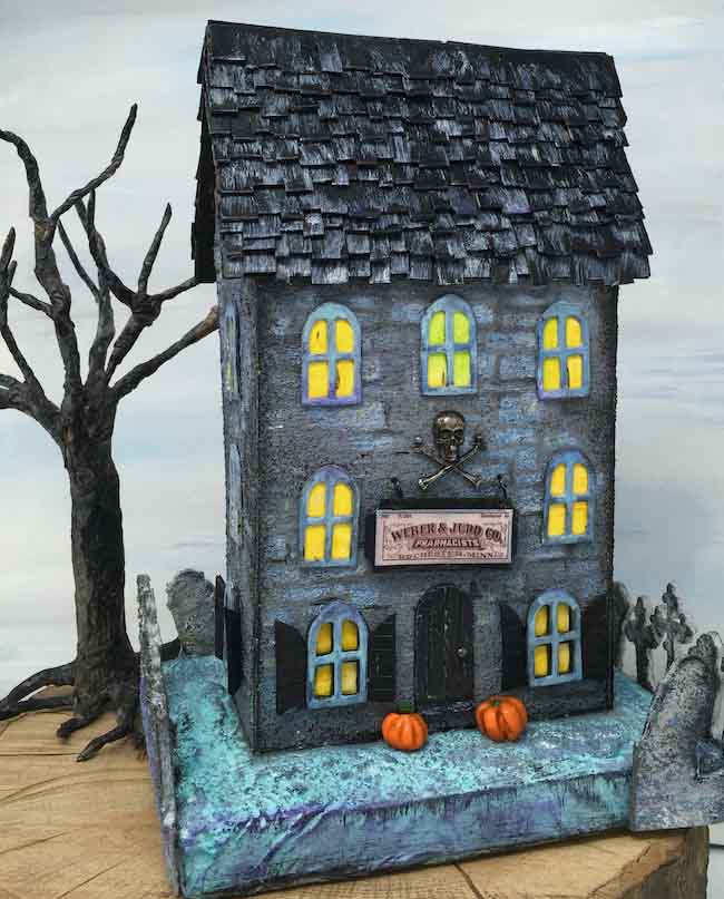 Cardboard Halloween house painted with blue and grey stones. Spooky paper mache tree beside the house. 